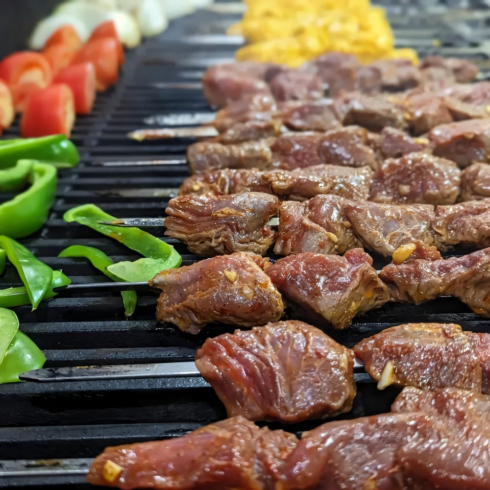 Marinated beef and chicken skewers with bell peppers, tomatoes, and onions grilling on a black grate.