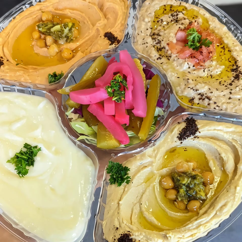 A Mazaj Mediterranean platter with four dips: two hummus, one baba ghanoush, and one garlic sauce, plus pickles.